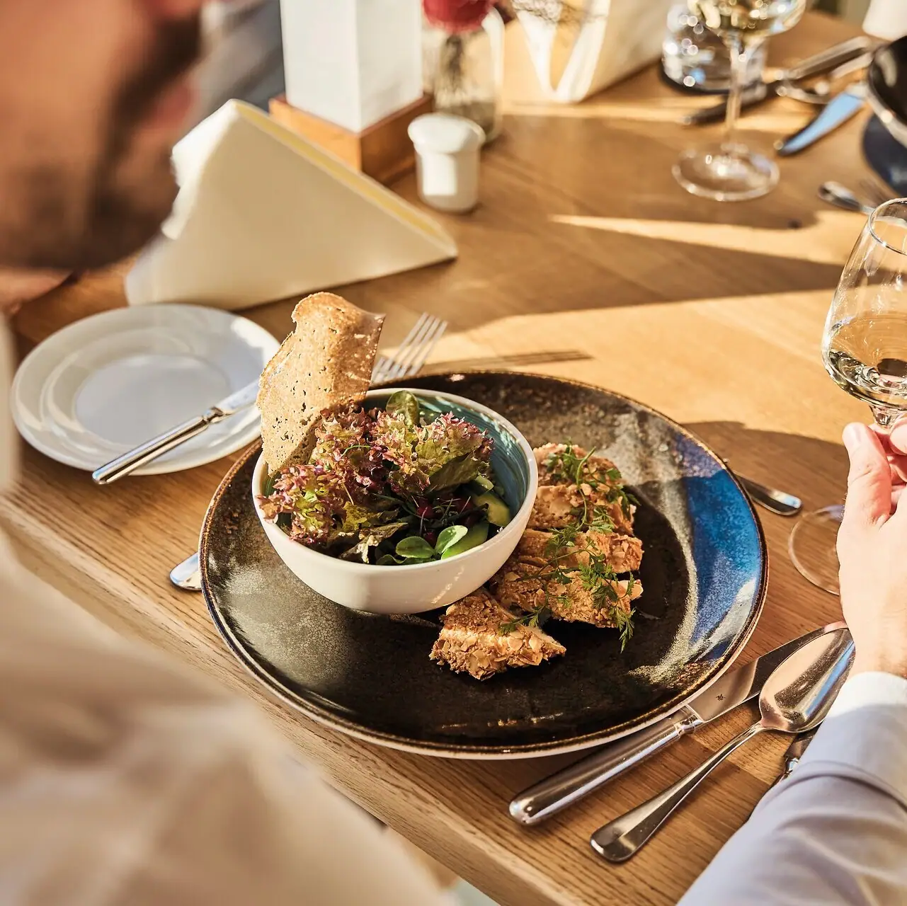 Ein Mann isst von einem Teller und hält ein Glas Wein. Auf dem Tisch stehen zwei Gerichte. Die Sonne scheint auf den Tisch.