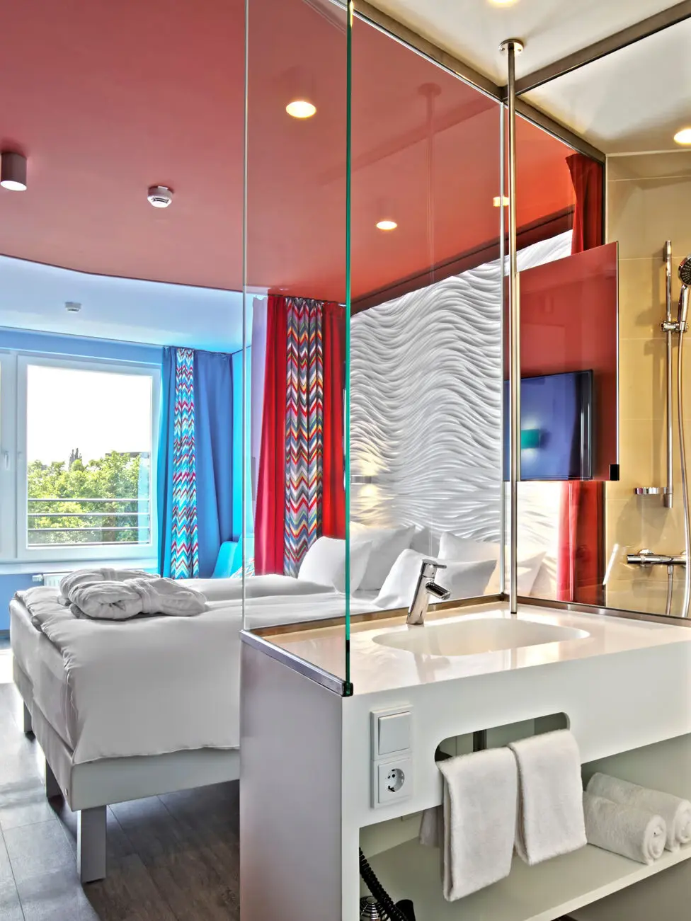 Ein Hotelzimmer mit einem Bad mit Waschbecken und Glaswand im Vordergrund und einem gemütlichen Bett und einem großen Fenster im Hintergrund.