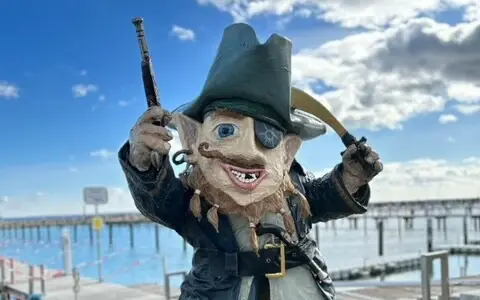 Digitale Schnitzeljagd Grömitz Ein Piratenfigur steht an einem Strand mit einem Steg im Hintergrund.