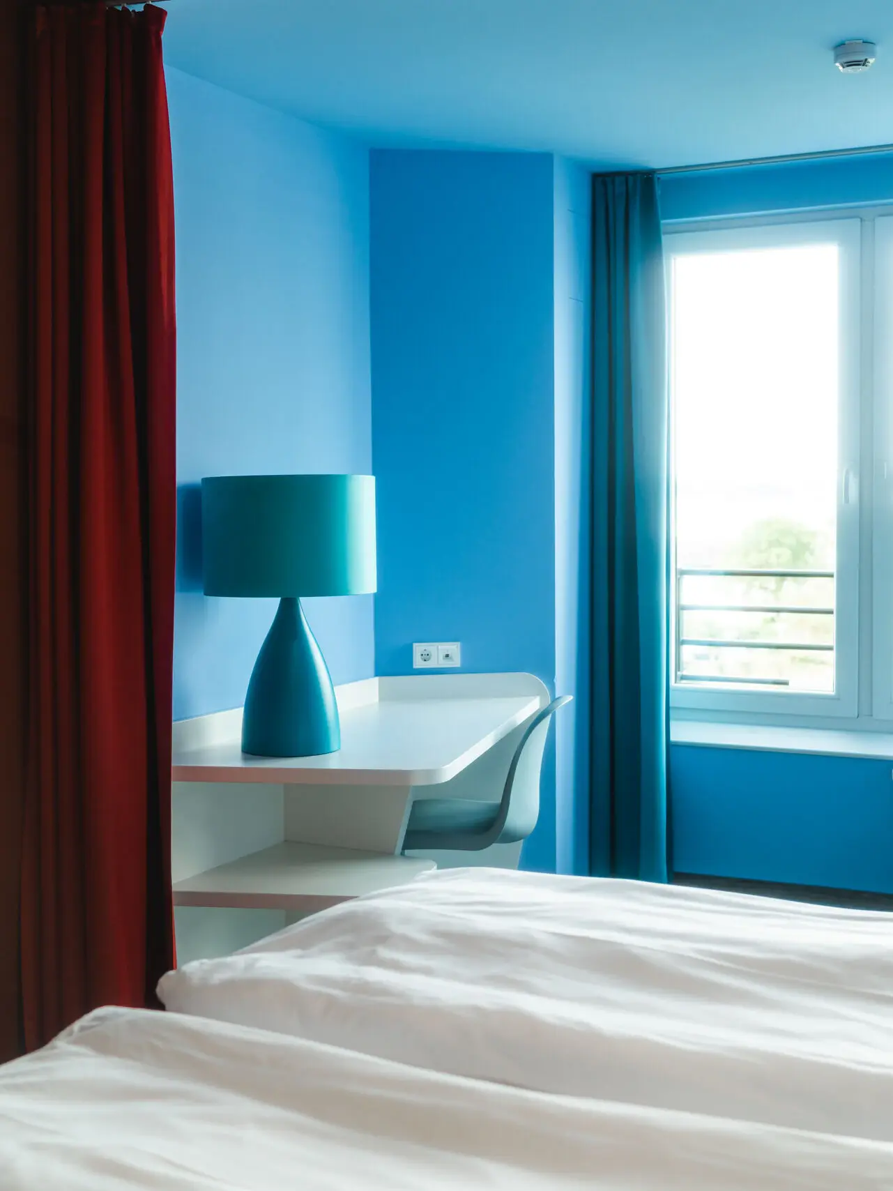 Ein Hotelzimmerauschnitt mit blauen Wänden. Hinter einer weißen Bettecke steht ein Schreibtisch mit einer blauen Lampe darauf.