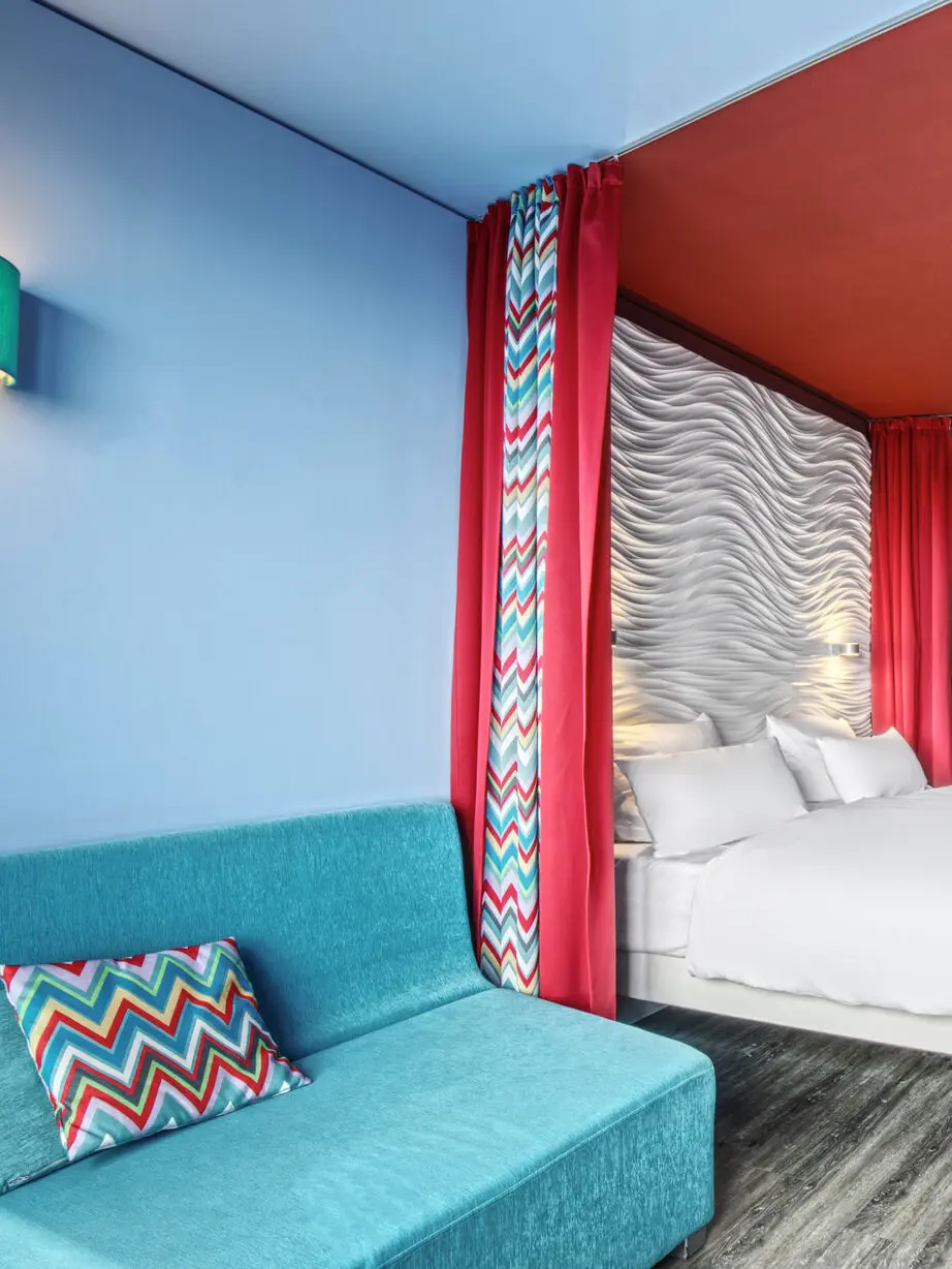 Ein Zimmerausschnitt mit grauem Teppich. Vor einer hellblauen Wand steht ein türkisfarbenes Sofa und dahinter vor einer roten Wand ein Bett mit welligem Kopfteil, das durch einen zurückgezogenen Vorhang vom vorderen Teil separiert werden kann.