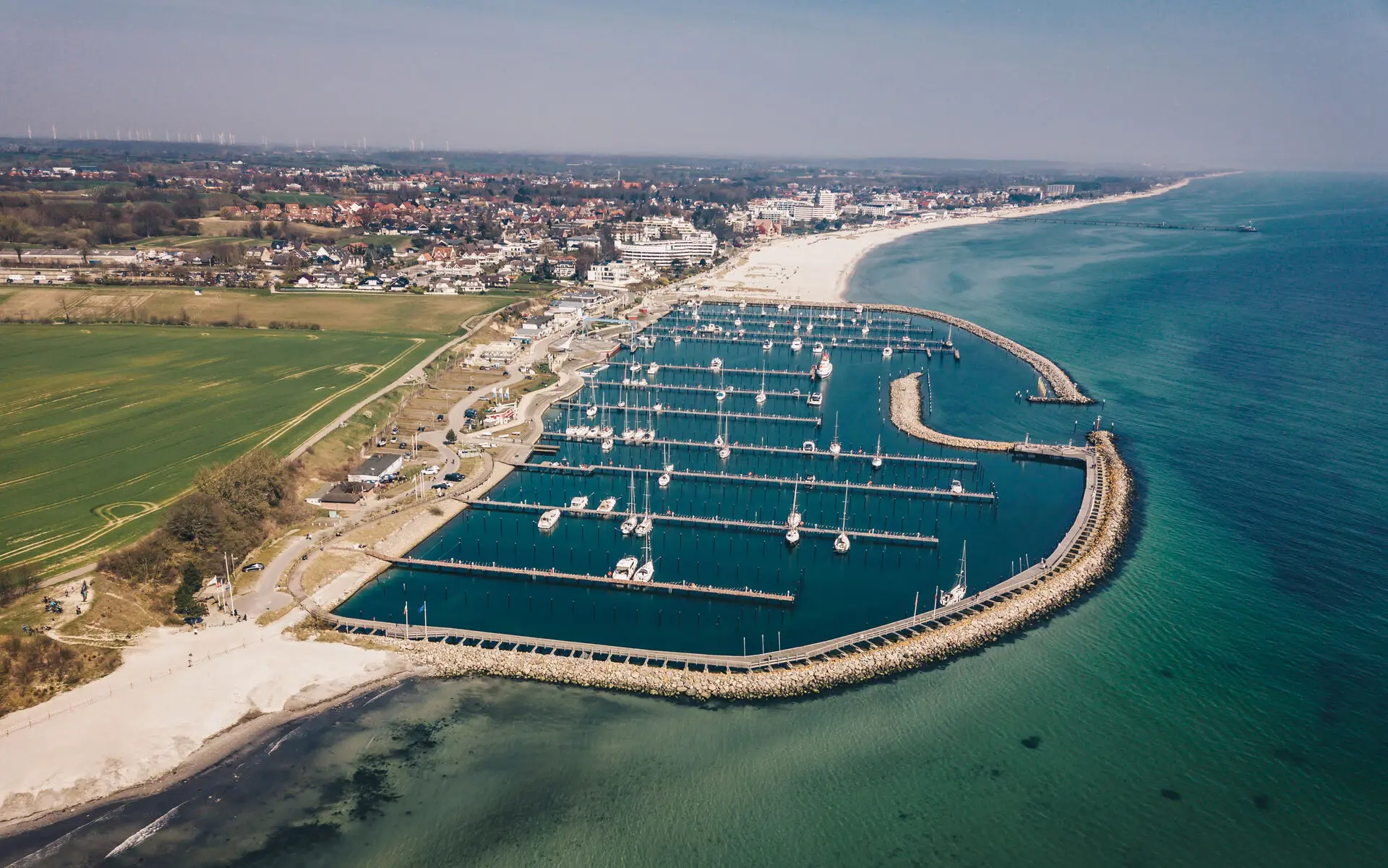 Eine Luftaufnahme eines Yachthafens, der an einen Strand und weite Wiesen angrenzt und in das Meer übergeht.