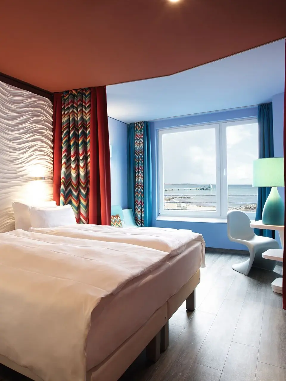Ein Hotelzimmer in gemütlichem Licht mit einem Holzboden und roter Decke. Das Bett ist durch kleine Lampen beleuchtet und im Hintergrund trennt ein zurückgezogener Vorhang den Schlafbereoch von einem dahinter stehenden Sofa und großen Fenster ab.