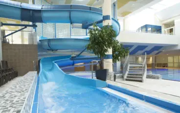 In einem hellgefliesten Badebereich schlängelt sich eine blaue Wasserrutsche hinunter in ein kleines Becken.