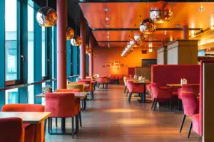 Ein warm beleuchtetes Restaurant mit Holzboden und roten Wänden. Entlang einer Fensterfront sowie im Rest des Raumes stehen rote Sitzgarnituren an Holztischen. Viele runde goldene Lampen hängen über den Tischen und tauchen den Raum in warmes Licht.