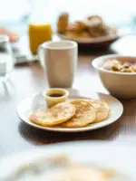 Auf einem Tisch stehen Teller mit Pancakes, Müslischalen und Kaffeebecher.