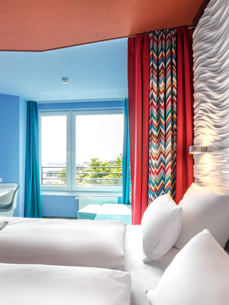 Ein helles Hotelzimmer, das durch einen Vorhang in den vorderen Schlafbereich mit einer roten Decke und einem gemütlichen weißen Bett und den dahinter liegenden Wohnbereich mit blauen Wänden, einem Sofa und Schreibtisch neben einem großen Fenster getrennt wird.