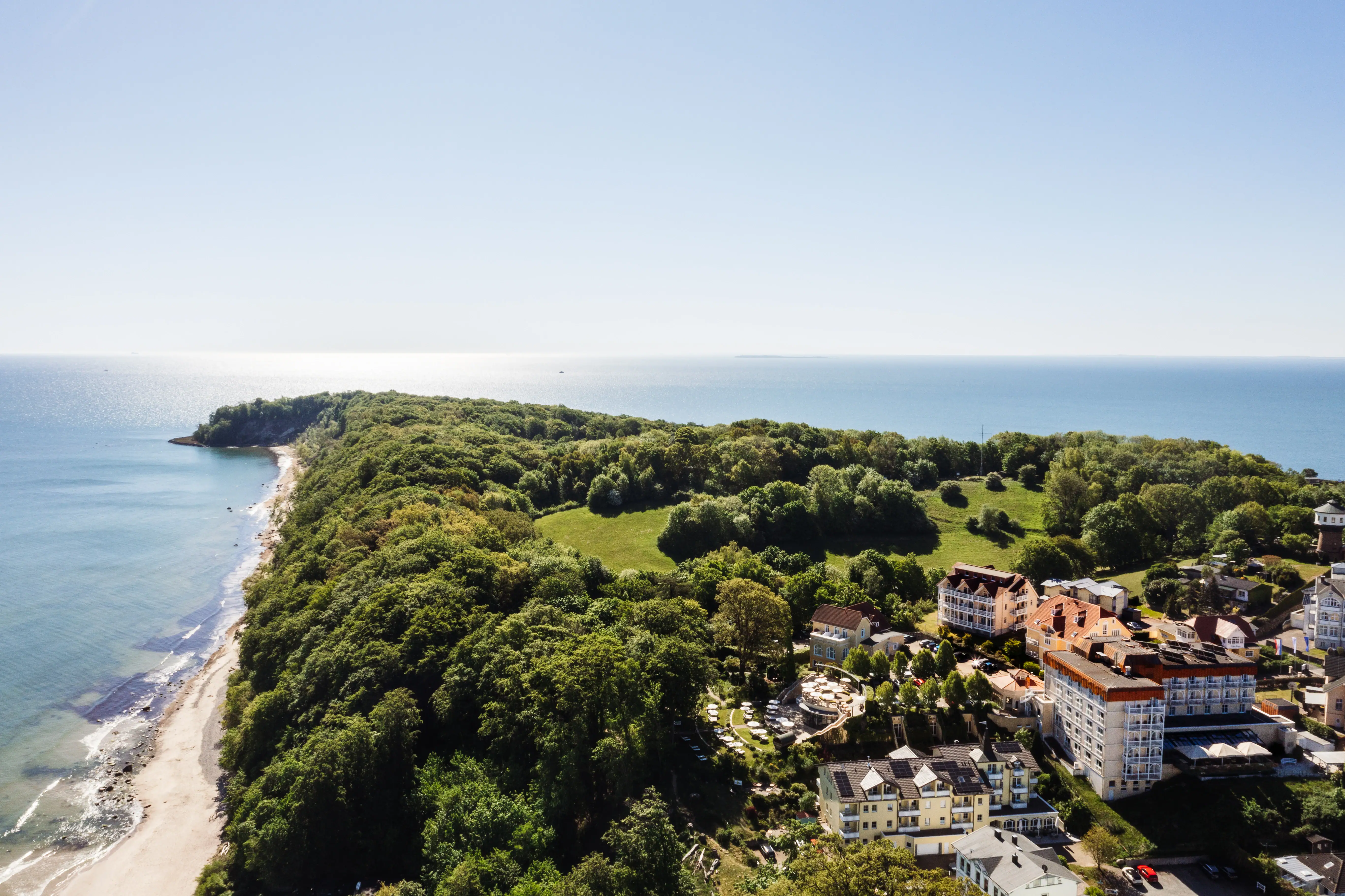 aja Nordperd & Villen Landschaft mit Bäumen und Gebäuden, im Hintergrund Küstenlinie und Meer.