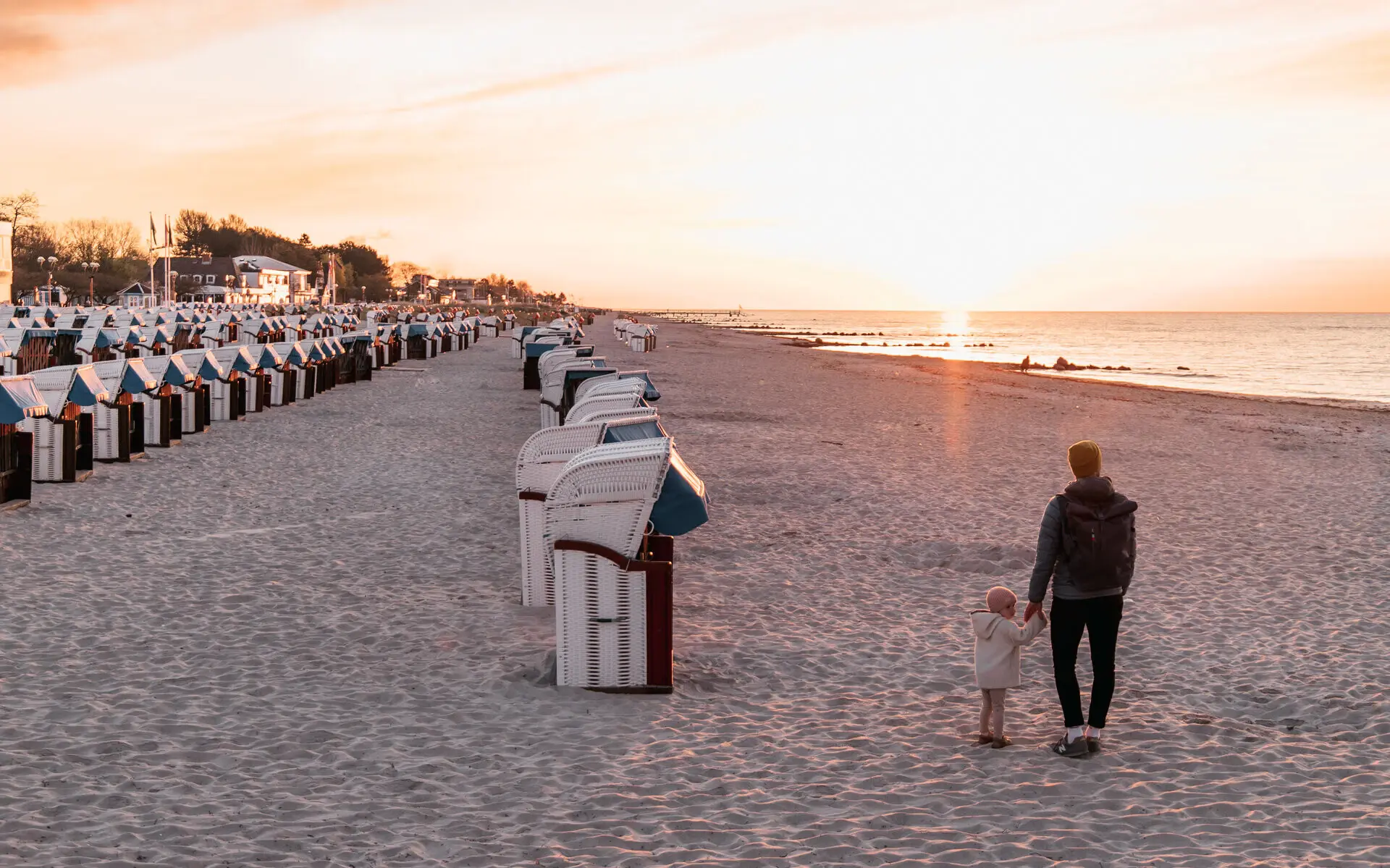 Ein Mann geht mit seiner kleinen Tochter an einem ruhigen Strand spazieren. Lange Reihen an Strandkörben führen den Strand entlang und in der ferne get die Sonne in warmen Farben über dem Wasser unter.