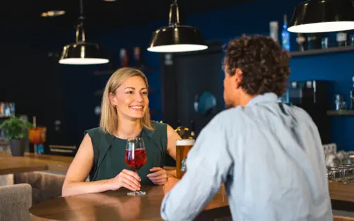 Ein Paar sitzt an einer Bar mit blauen Wänden und stößt lachend mit Getränken an. Ein Paar sitzt an einer Bar mit blauen Wänden und stößt lachend mit Getränken an.