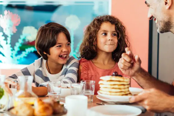 Frühstück im aja Warnemünde Zwei Kinder sitzen mit dem Vater im Familienbereich an einem Tisch mit Pfannkuchen, Getränken und Brötchen.