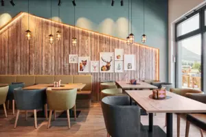 Gemütlicher Essbereich mit Holzwand und dekorativen Tierbildern. Hängelampen und warmes Licht schaffen eine einladende Atmosphäre und verschiedne Sitzmöglichkeiten bieten Platz für Gäste. Durch ein Fenster sieht man eine Außenlandschaft mit Bergen.