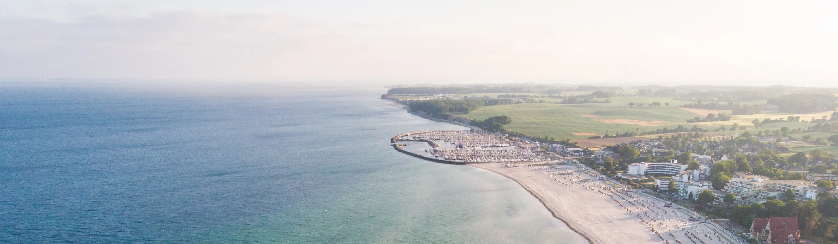 Luftansicht eines Küstenbereichs mit blauem Meer, einem kleinen Yachthafen und grünen Wäldern in der Sonne.