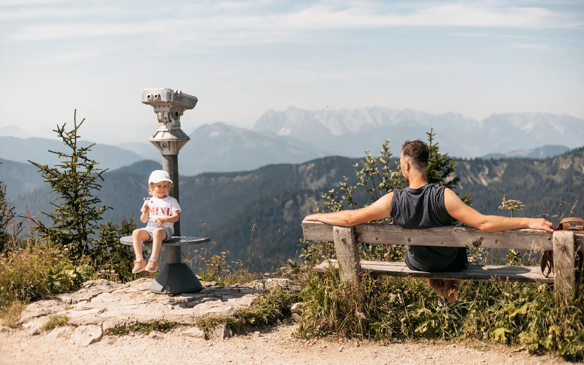 Hochfelln Ein Mann sitzt auf einer Bank in den bergen und schaut zu einem kleinen Mädchen hinüber, welches lächelnd auf der Plattform eines Fernglases sitzt. Im Hintergrund erstreckt sich eine Berglandschaft und die Sonne scheint warm vom Himmel.