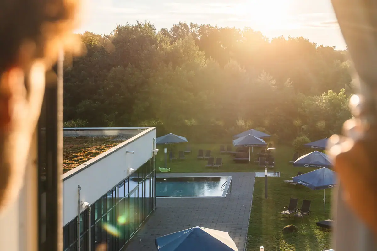 Ausblick vom Zimmer im aja Bad Saarow Eine Person blickt aus einem Fenster auf einen Pool mit Sonnenschirmen.
