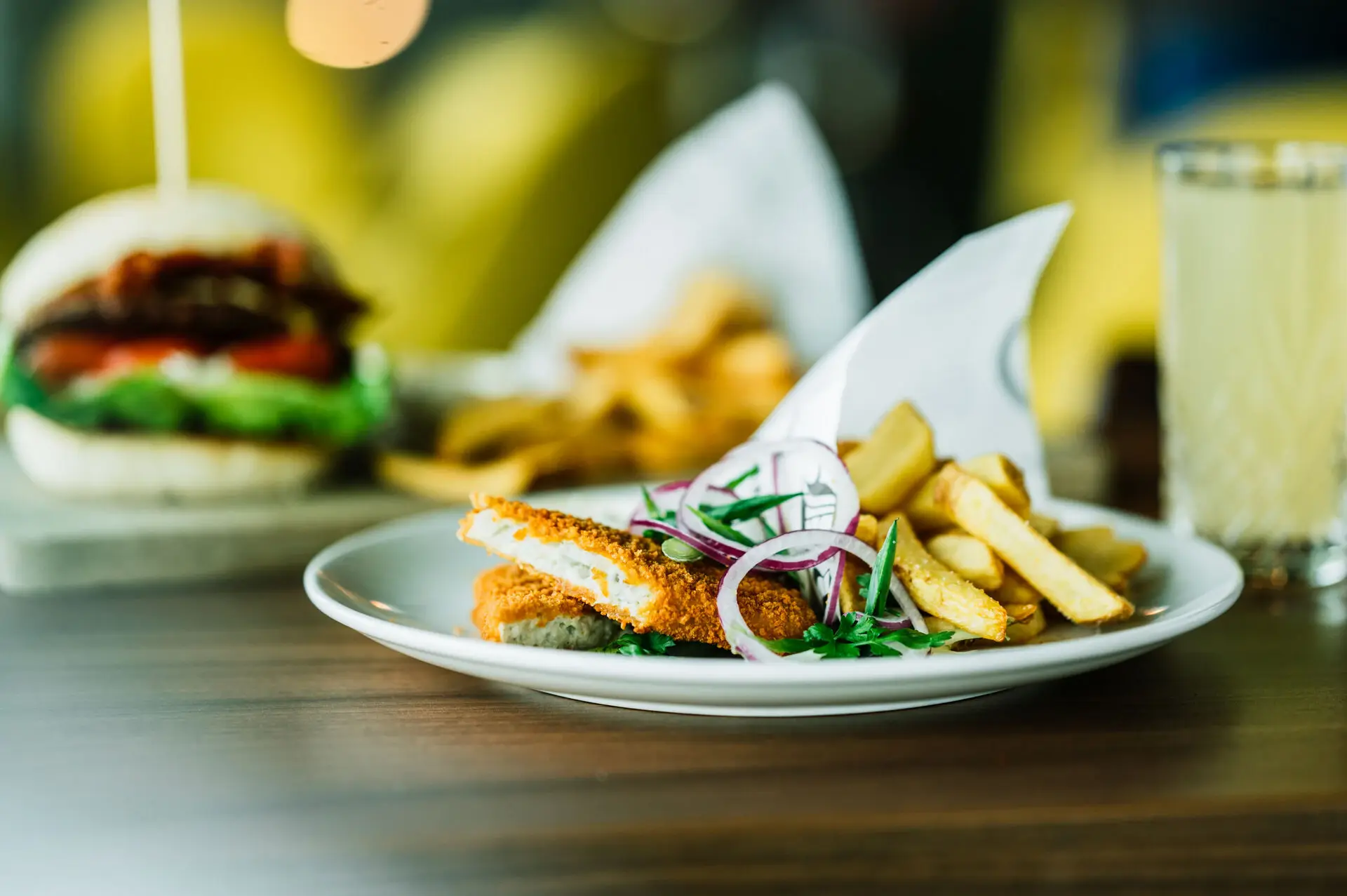 Ein Teller mit knusprigem paniertem Fisch, garniert mit roten Zwiebeln und frischen Kräutern, serviert mit goldgelben Pommes frites. Im Hintergrund ein Burger und ein Glas Getränk.