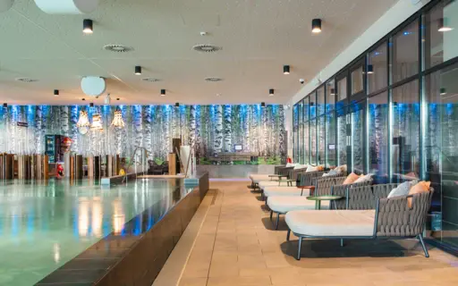 Der Innenpool im aja Bad Saarow mit türkis beleuchtetem Wasser und umstehenden Loungebetten. Der Innenpool im aja Bad Saarow mit türkis beleuchtetem Wasser und umstehenden Loungebetten.