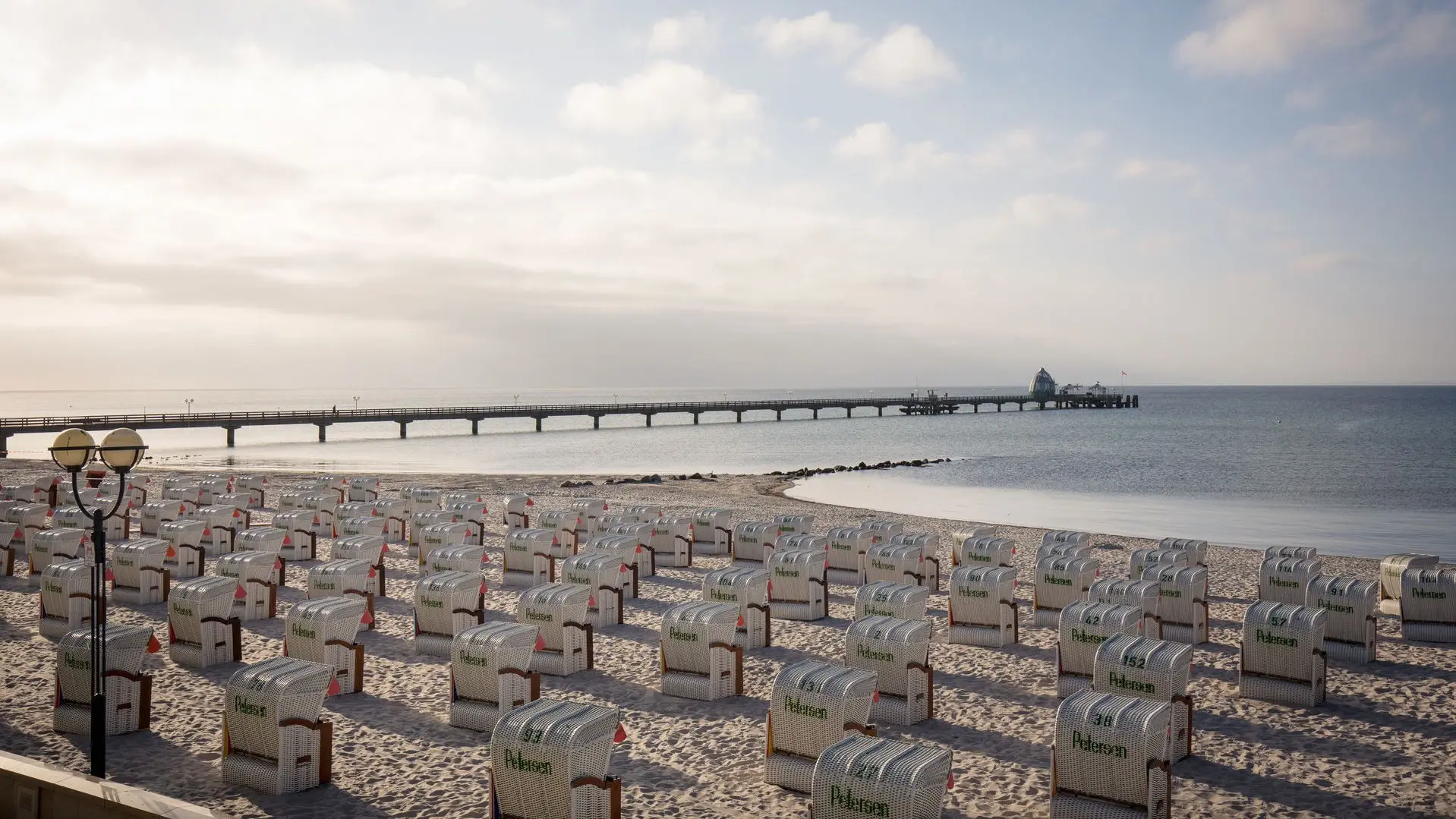An einem leeren Strand stehen viele Strandkörbe in Richtung Meer aufgereiht. Ein langer Steg führt auf das Wasser hinaus und die Morgensonne scheint warm vom Himmel.