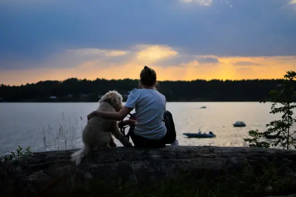 Eine Frau und ein Hund sitzen auf einem Baumstamm am Wasser.