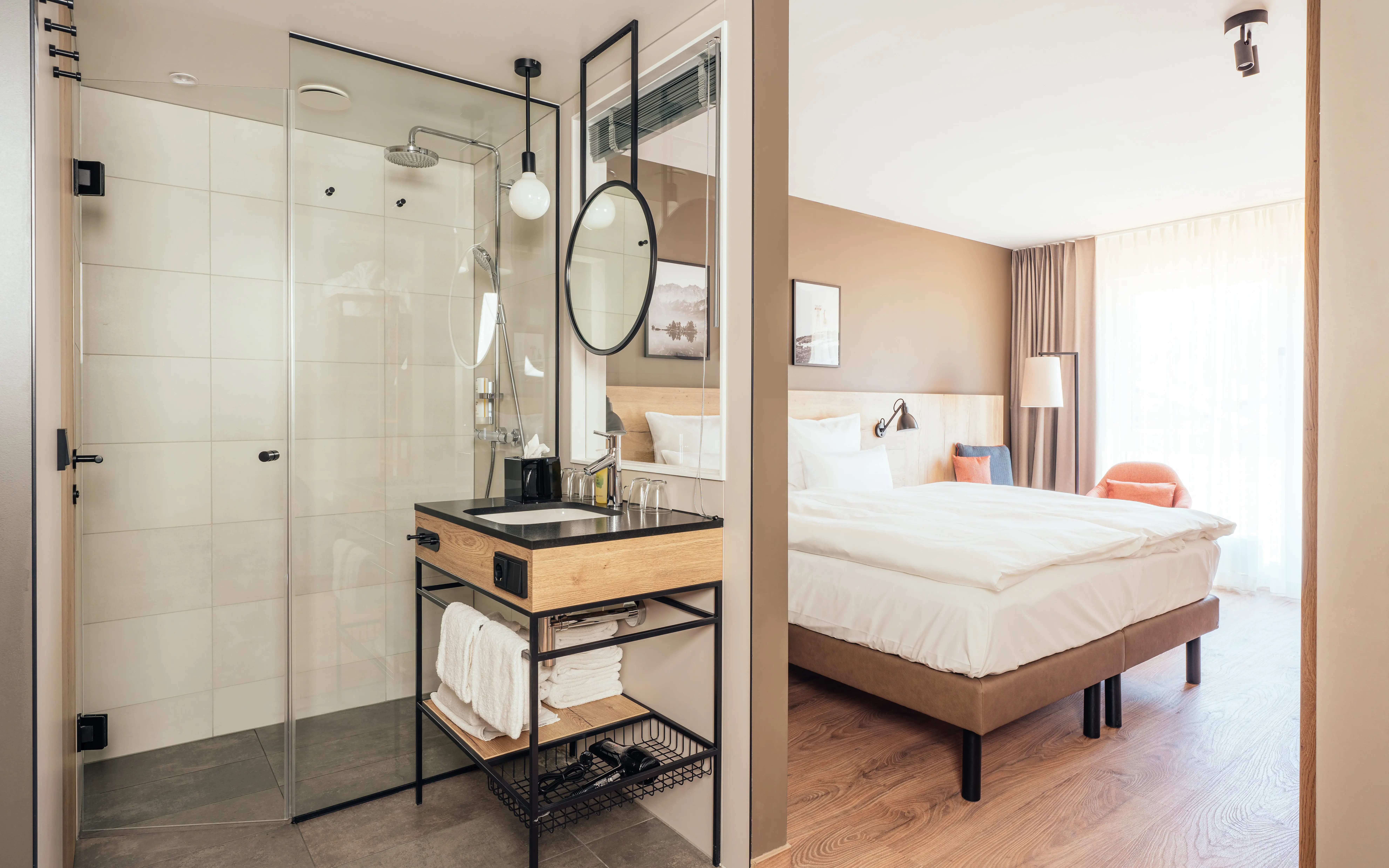 Modernes Hotelzimmer mit offenem Badezimmerkonzept. Eine Glasdusche und ein stilvolles Waschbecken mit Holz- und Metallakzenten befinden sich direkt neben dem Schlafbereich. Das Zimmer ist in warmen, natürlichen Tönen gehalten, mit einem großen Bett, gemütlichem Sessel und einer Fensterfront, die viel Licht hereinlässt.