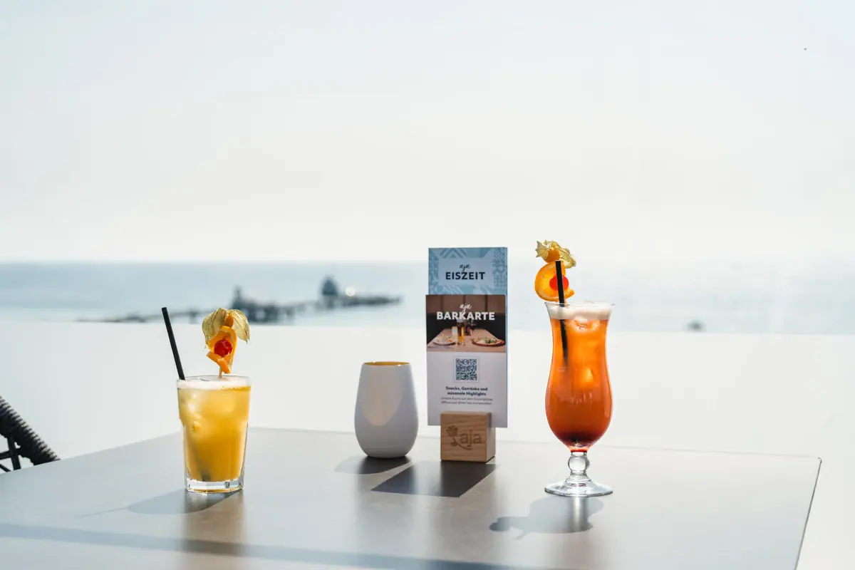 Cocktails Rooftop Bar aja Grömitz Zwei Cocktails auf einem Tisch in der aja Rooftop Bar Grömitz mit Meerblick und der Seebrücke im Hintergrund.