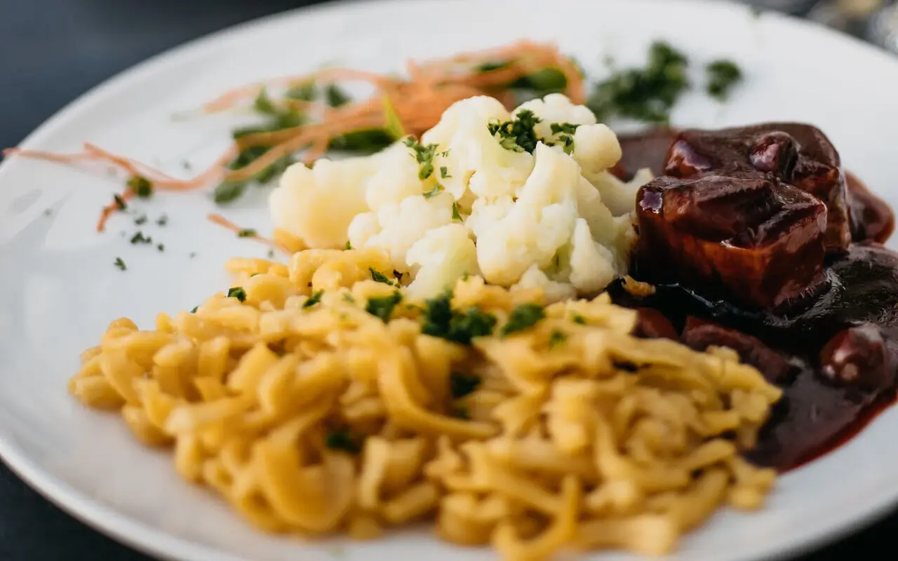 Abendessen aja Ruhpolding Auf einem weißen Teller ist ein Abendessen aus Spätzle, Blumenkohl und Fleisch in einer braunen Soße angerichtet.