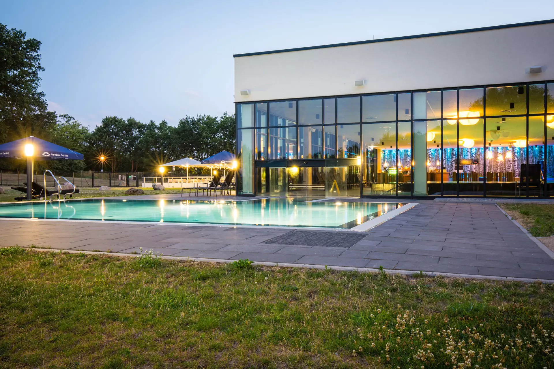 Ein bei Abend beleuchteter Außenpool mit umliegender Rasenfläche, der an einen SPA-Bereich anschließt.