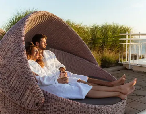 Wellness Ein Mann und eine Frau in weißen Bademänteln sitzen auf einer gemütlichen runden Liege.