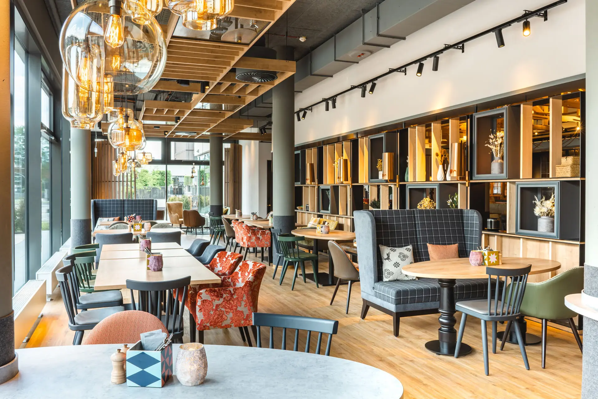 Heller, stilvoller Restaurantbereich mit großen Fenstern, die viel Tageslicht hereinlassen.Bunte Stühle und gemütliche Nischen schaffen eine einladende Atmosphäre. Die dekorativen Regale an der Wand sind mit Trockenblumen und anderen Akzenten bestückt, die dem Raum einen warmen, modernen Charme verleihen.
