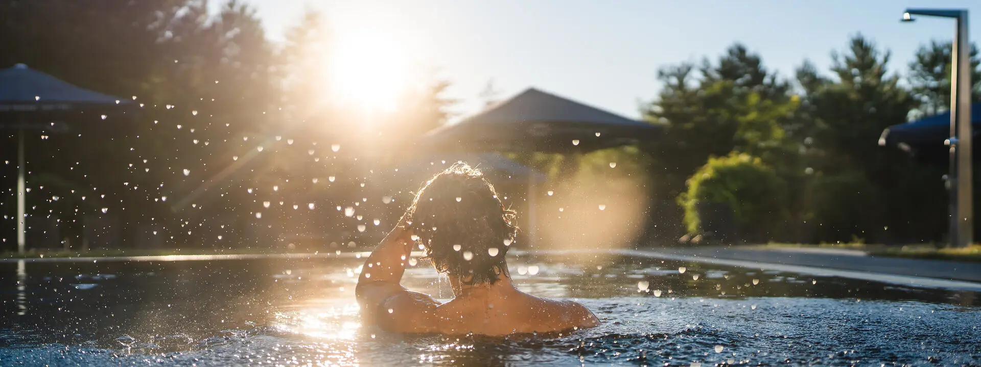 Außenpool aja Bad Saarow Ein Mann schwimmt im Außenpool vom aja Bad Saarow und wischt sich mit einer Hand die Haare aus dem Gesicht während Wassertropfen durch die Luft spritzen. Am Himmel leuchtet die Morgensonne.
