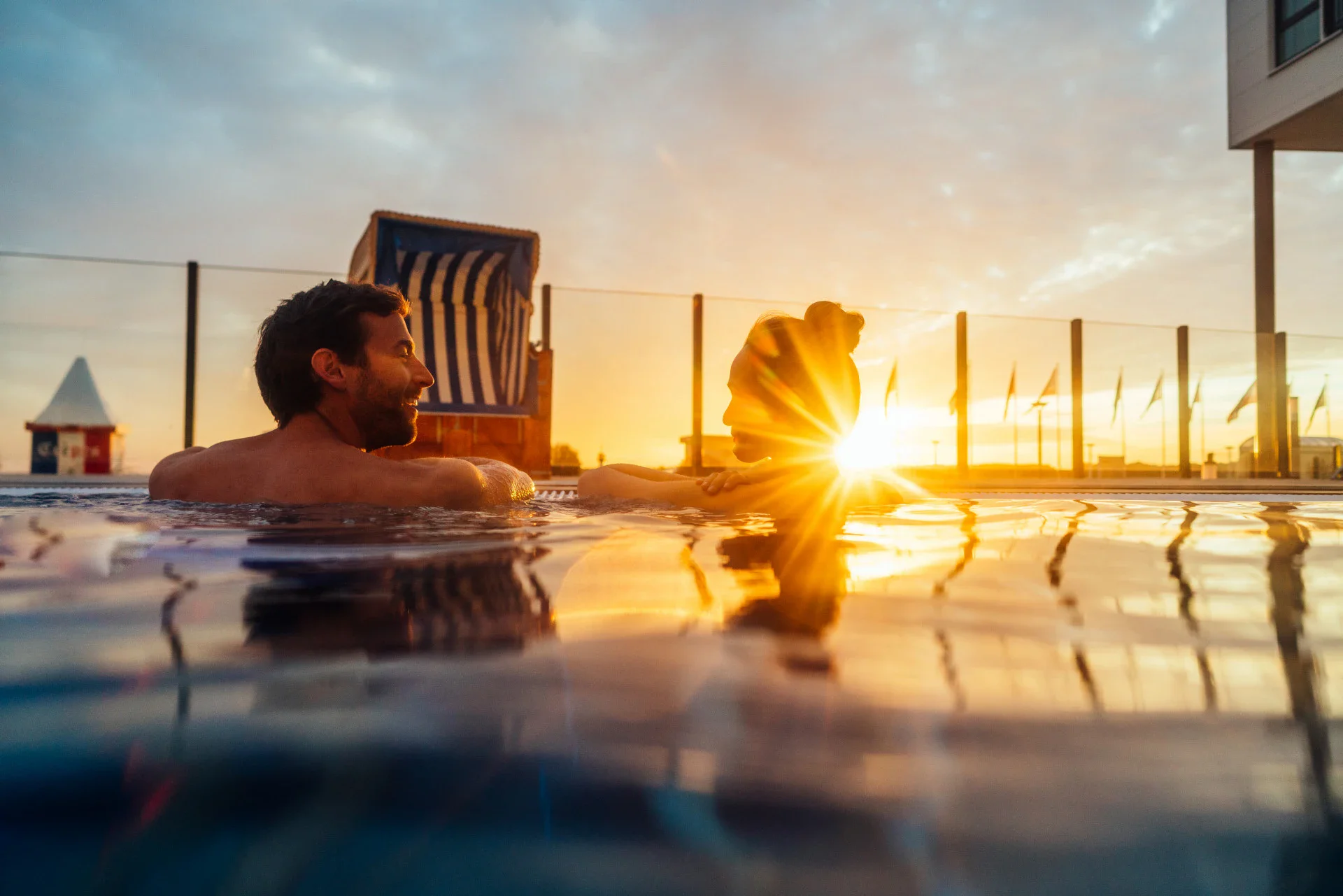 Ein Mann und eine Frau genießen den Sonnenuntergang in einem Außenpool, während das warme Licht der untergehenden Sonne die Szene stimmungsvoll beleuchtet. Im Hintergrund ist ein Strandkorb zu sehen.