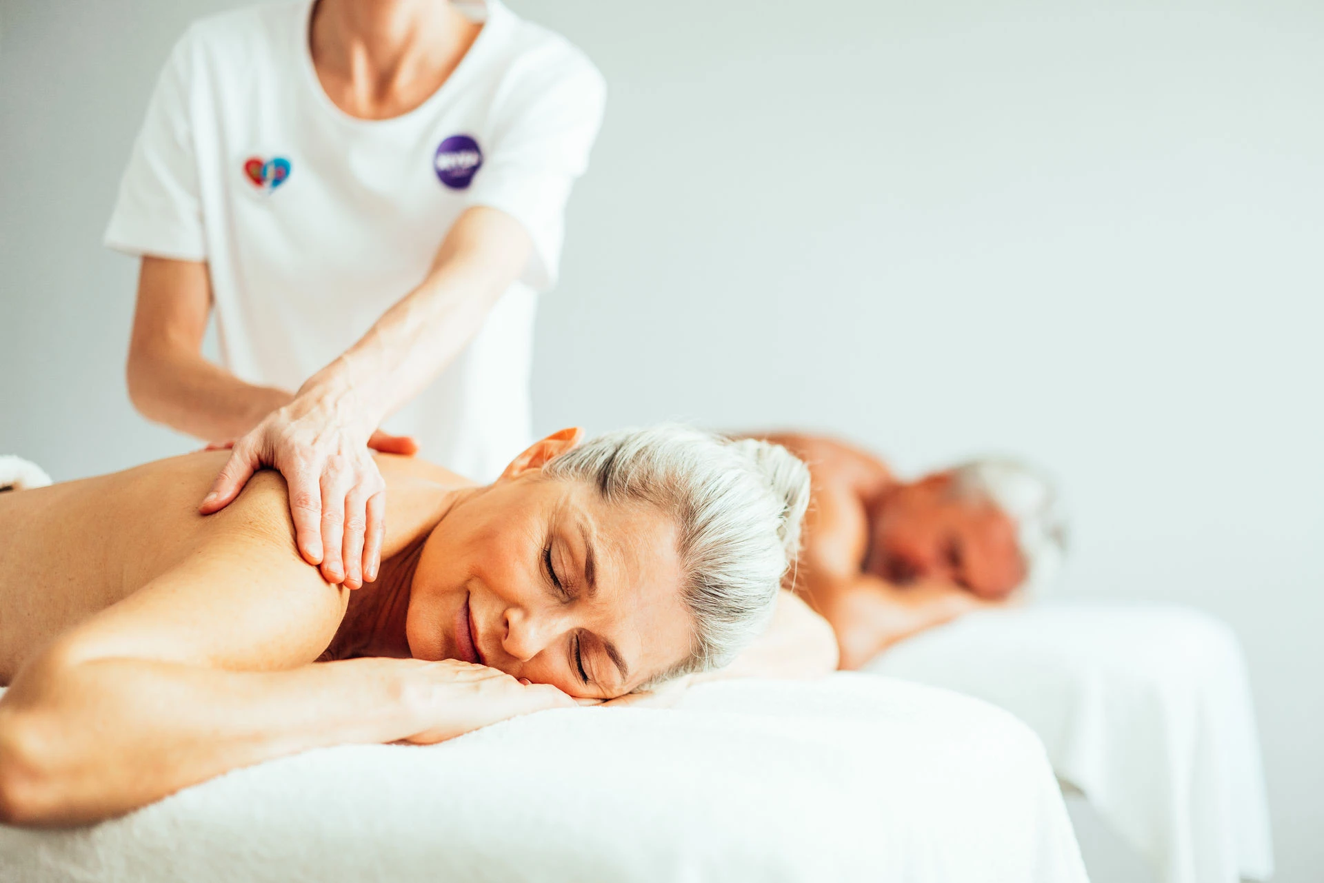 Eine Frau genießt eine entspannende Rückenmassage und lächelt mit geschlossenen Augen, während ein Massagetherapeut sanft ihre Schultern bearbeitet. Im Hintergrund liegt ein Mann ebenfalls entspannt auf einer Massageliege.