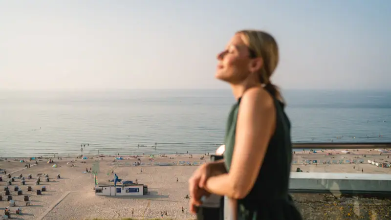 Zimmer im aja Warnemünde Eine Frau steht auf einem Geländer mit Blick auf einen Strand.