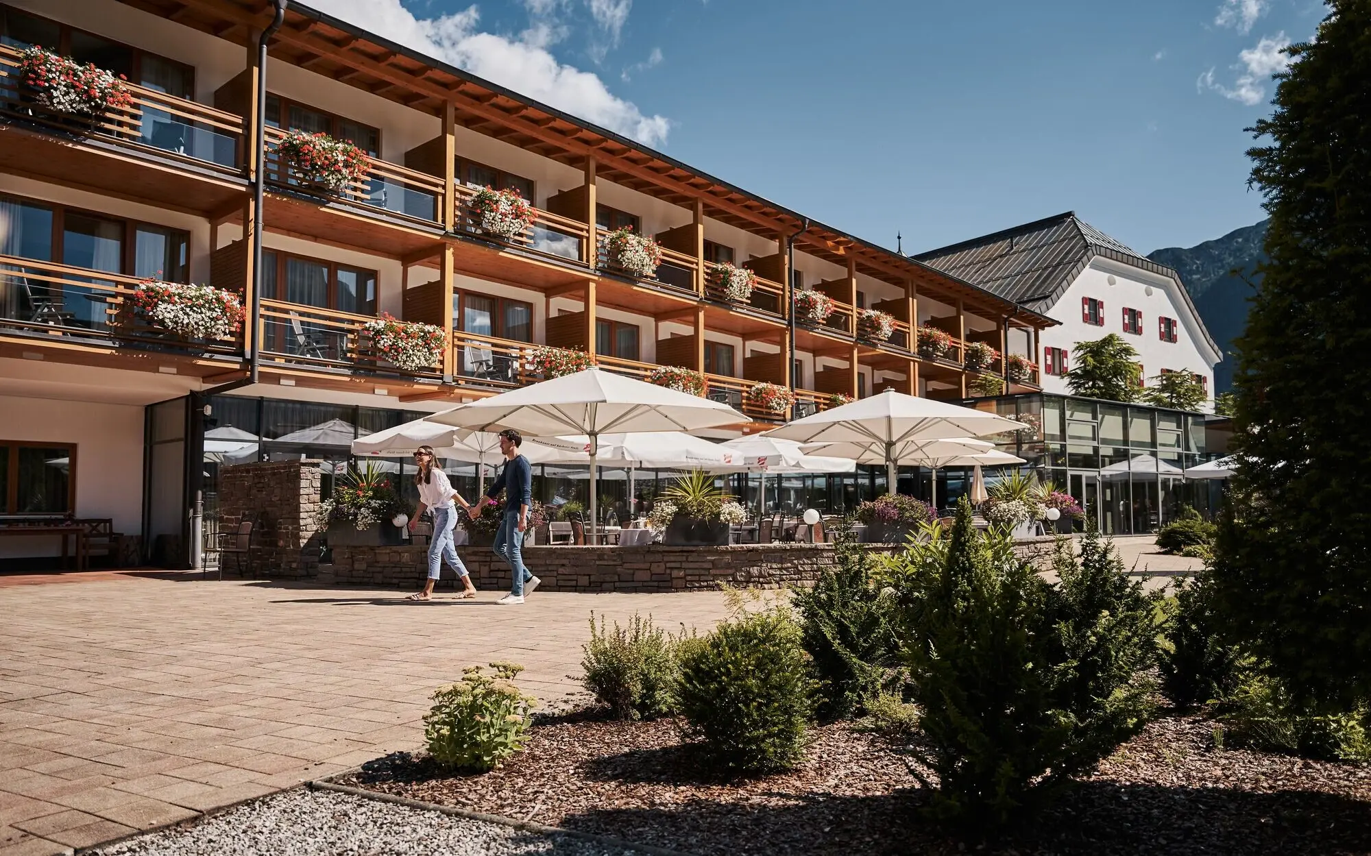 Gastgarten aja Fürstenhaus am Achensee Ein Paar spaziert vor einem Gebäude mit einer Außenterrasse und großen Sonnenschirmen.