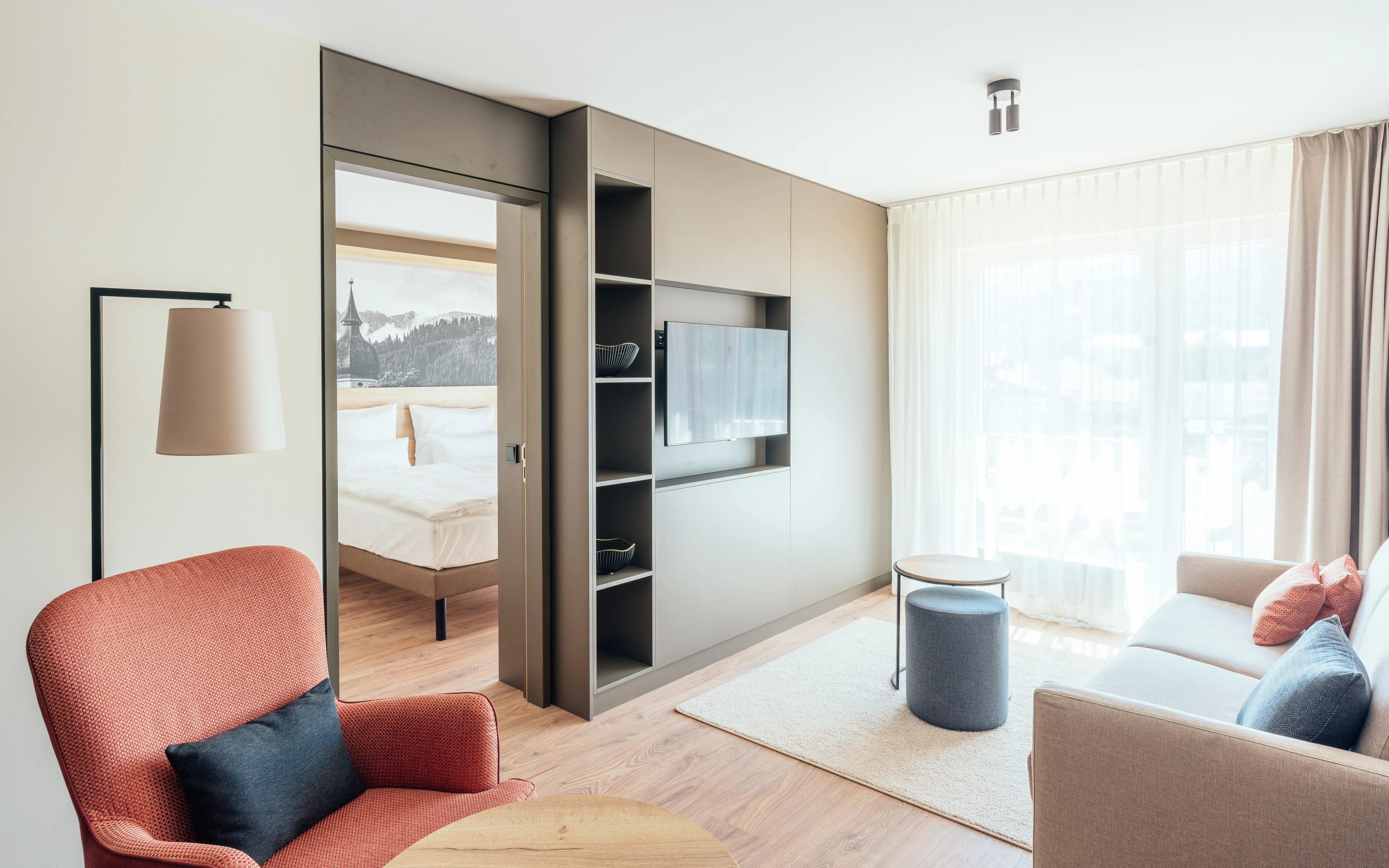 Modernes Hotelzimmer mit separatem Wohn- und Schlafbereich. Der Wohnbereich ist mit einem gemütlichen Sessel, einem Sofa und einem Flachbildfernseher ausgestattet. Eine große Fensterfront lässt viel Tageslicht herein. Im Hintergrund ist das Schlafzimmer mit einem großen Bett sichtbar, gestaltet in warmen Farben und minimalistischer Einrichtung.