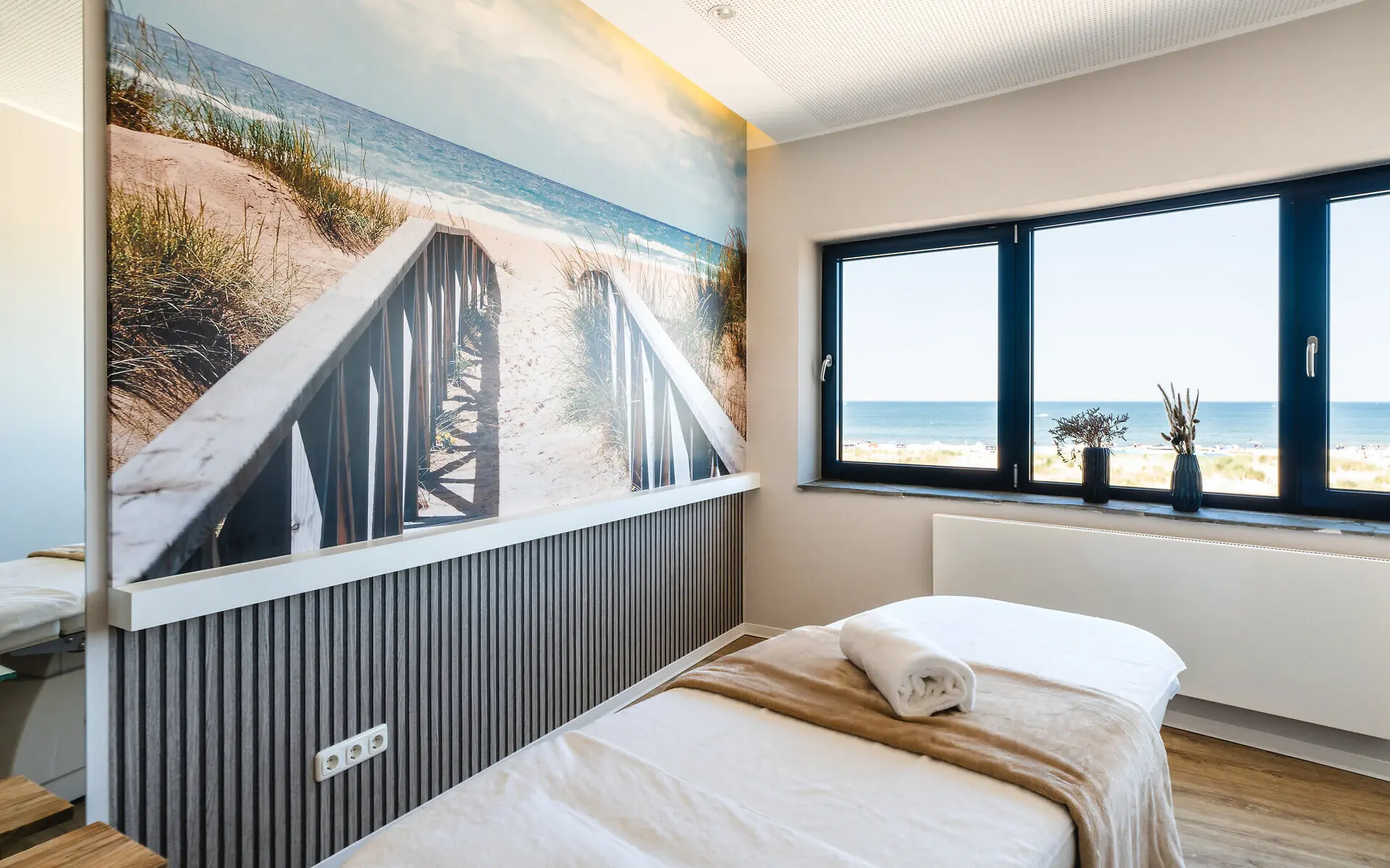 Anwendungsraum NIVEA Haus Warnemünde Vor einem großen Bild von einem Ostseezugang steht eine Behandlungsliege mit Blick auf die Ostsee durch ein großes Fenster, welches den Raum mit sonnigem Tageslicht durchflutet.