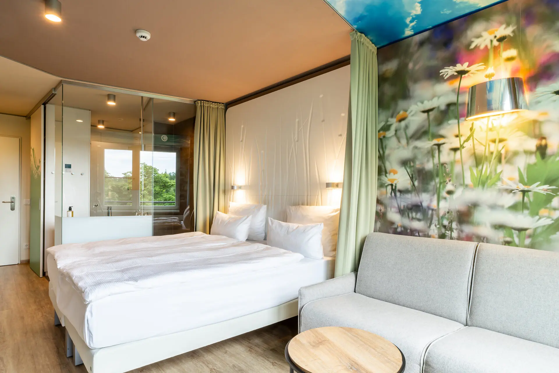Hotelzimmer im aja Bad Saarow Ein Hotelzimmer mit gemütlichem Bett und warmem Licht. Eine Sitzecke lädt zum Entspannen ein und die Tapeten sind mit einer realitätsgetreuen Blumenwiesentapete verziert.