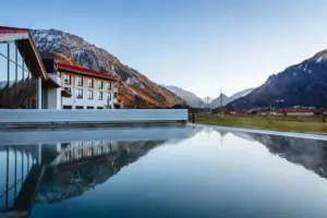 Außenansicht eines beheizten Außenpools mit klarem Wasser und sanftem Dampf, eingebettet in eine malerische Winterlandschaft. Im Hintergrund steht ein modernes Hotelgebäude mit rotem Dach, umgeben von schneebedeckten Bergen und Wiesen.