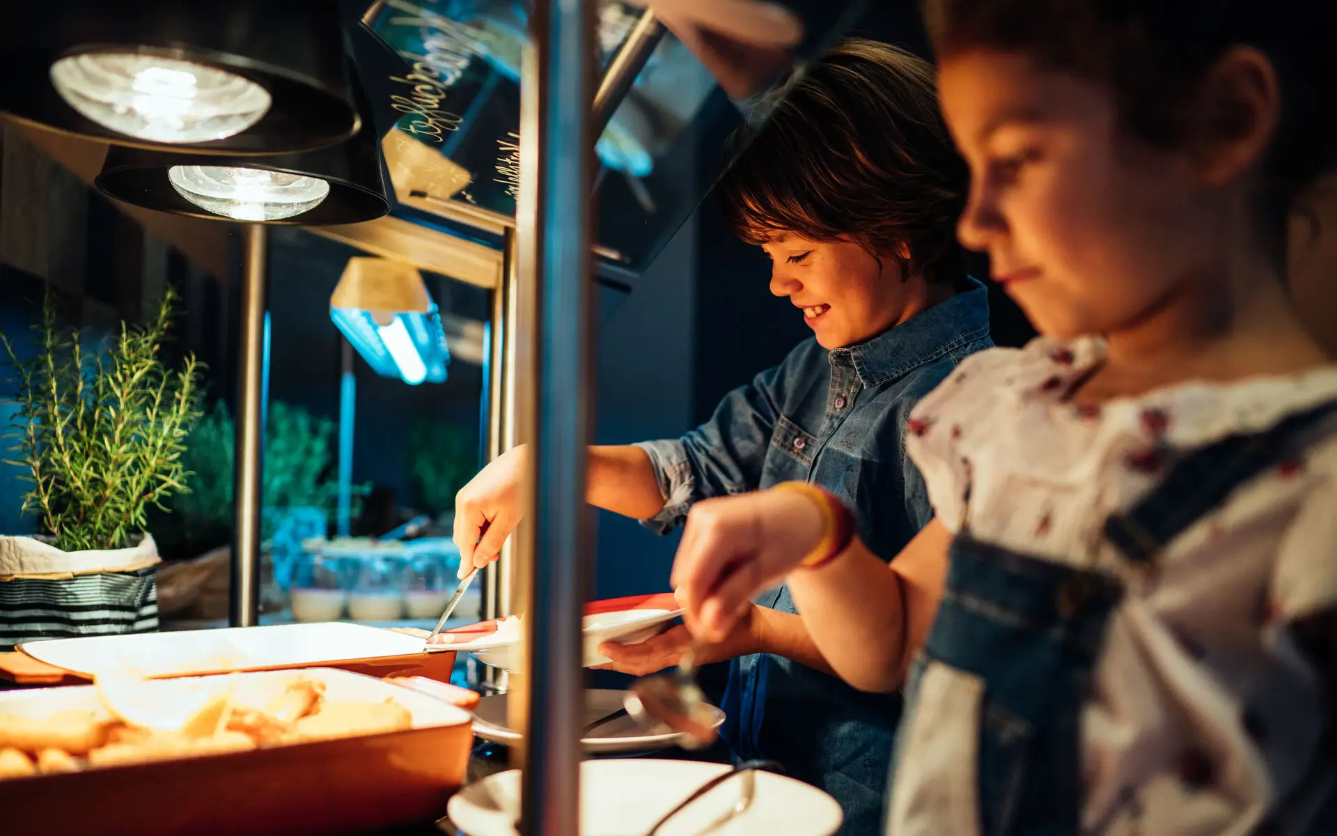 Kinderbuffet Zwei Kinder stehen an einem Buffet mit warmen Speisen und füllen sich Essen auf ihre Teller auf.