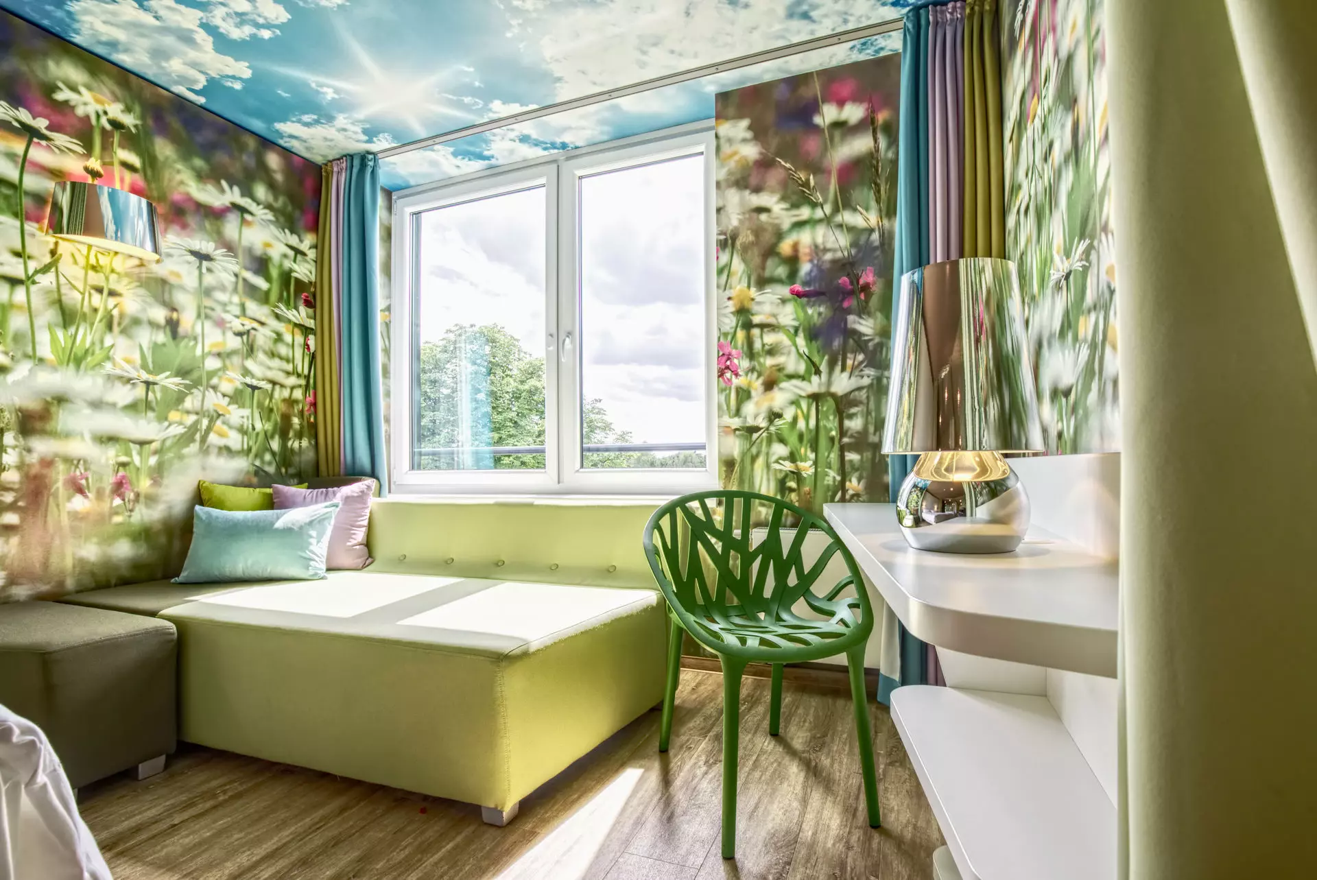 Ein Hotelzimmerausschnitt mit Blumenwiesetapete und einem kleinen Sofa mit bunten Kissen vor einem Fenster, das den Raum mit Licht füllt.