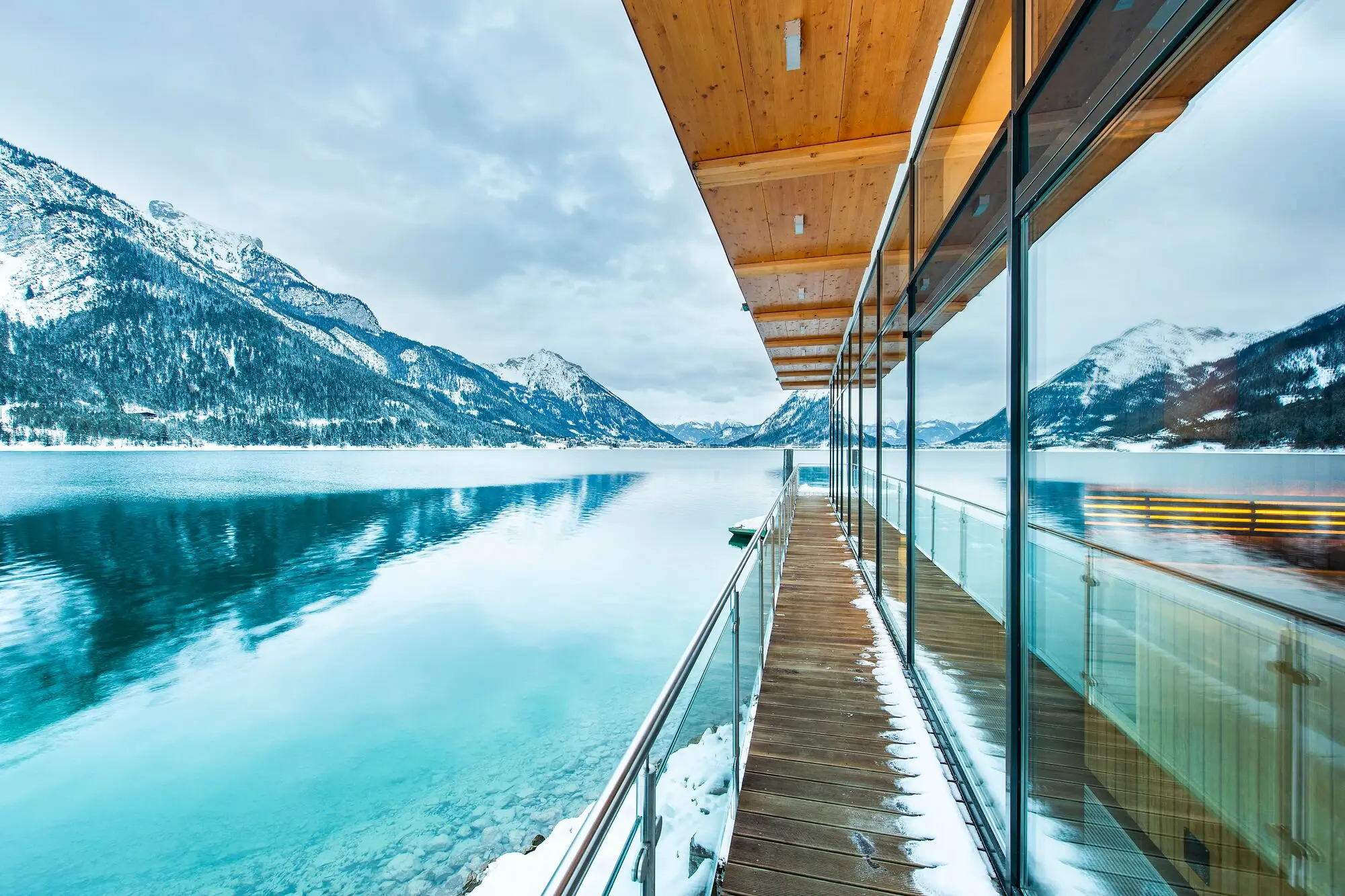aja Achensee Glaswand mit Holzdeck mit Blick auf einen See, umgeben von Bergen und Wolken.