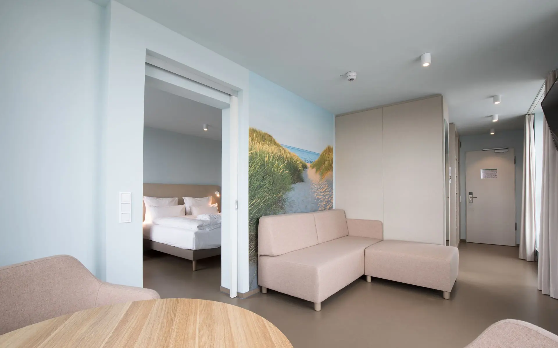 Ein Wohnbereich in einem Hotelzimmer mit hellem Sofa vor einer Wand mit einem Strandzugangaufdruck sowie einem runden Tisch mit zwei Stühlen. Eine Tür führt in das daneben liegende Schlafzimmer mit großem Bett.