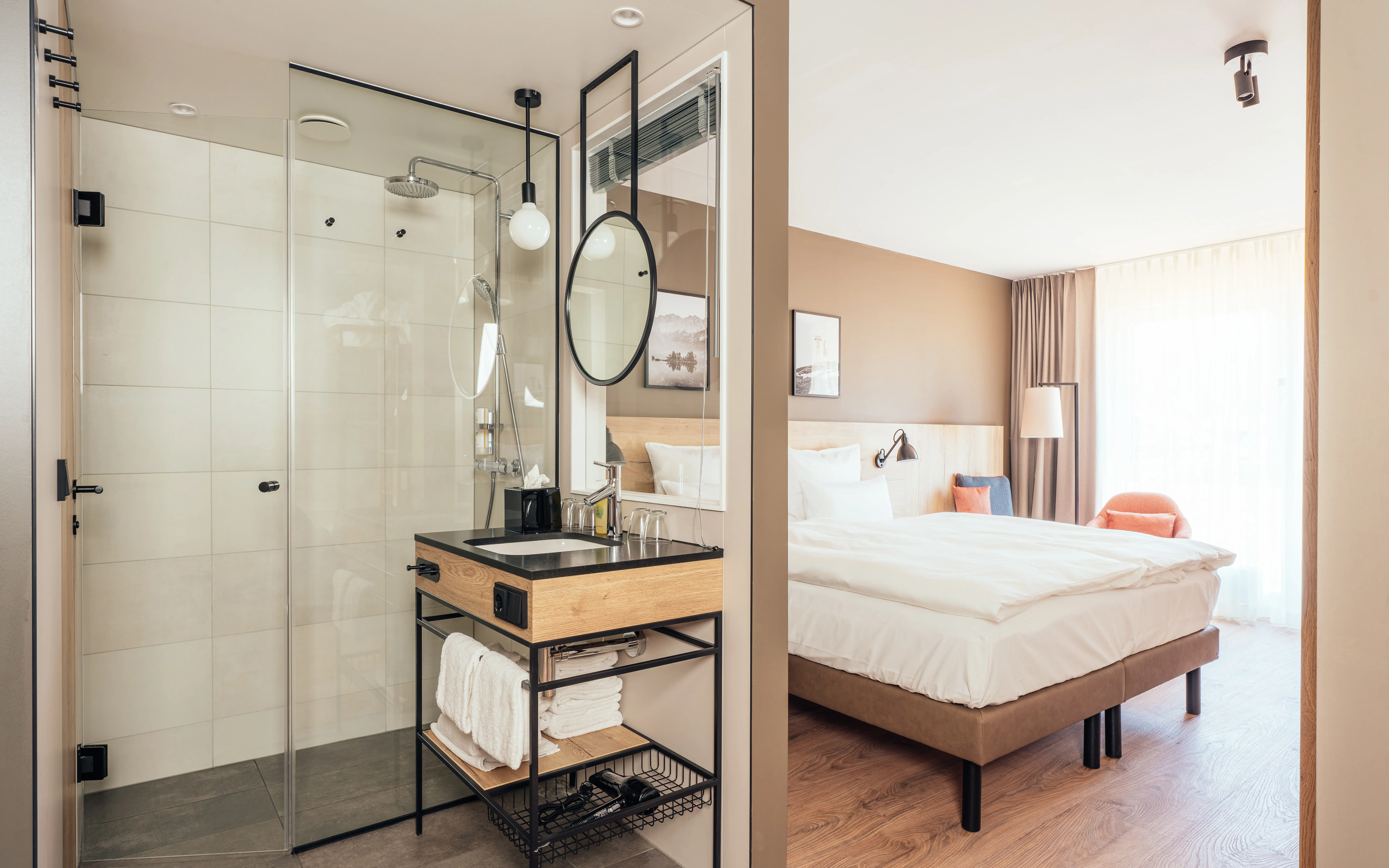 Modernes Hotelzimmer mit offenem Badezimmerkonzept. Eine Glasdusche und ein stilvolles Waschbecken mit Holz- und Metallakzenten befinden sich direkt neben dem Schlafbereich. Das Zimmer ist in warmen, natürlichen Tönen gehalten, mit einem großen Bett, gemütlichem Sessel und einer Fensterfront, die viel Licht hereinlässt.