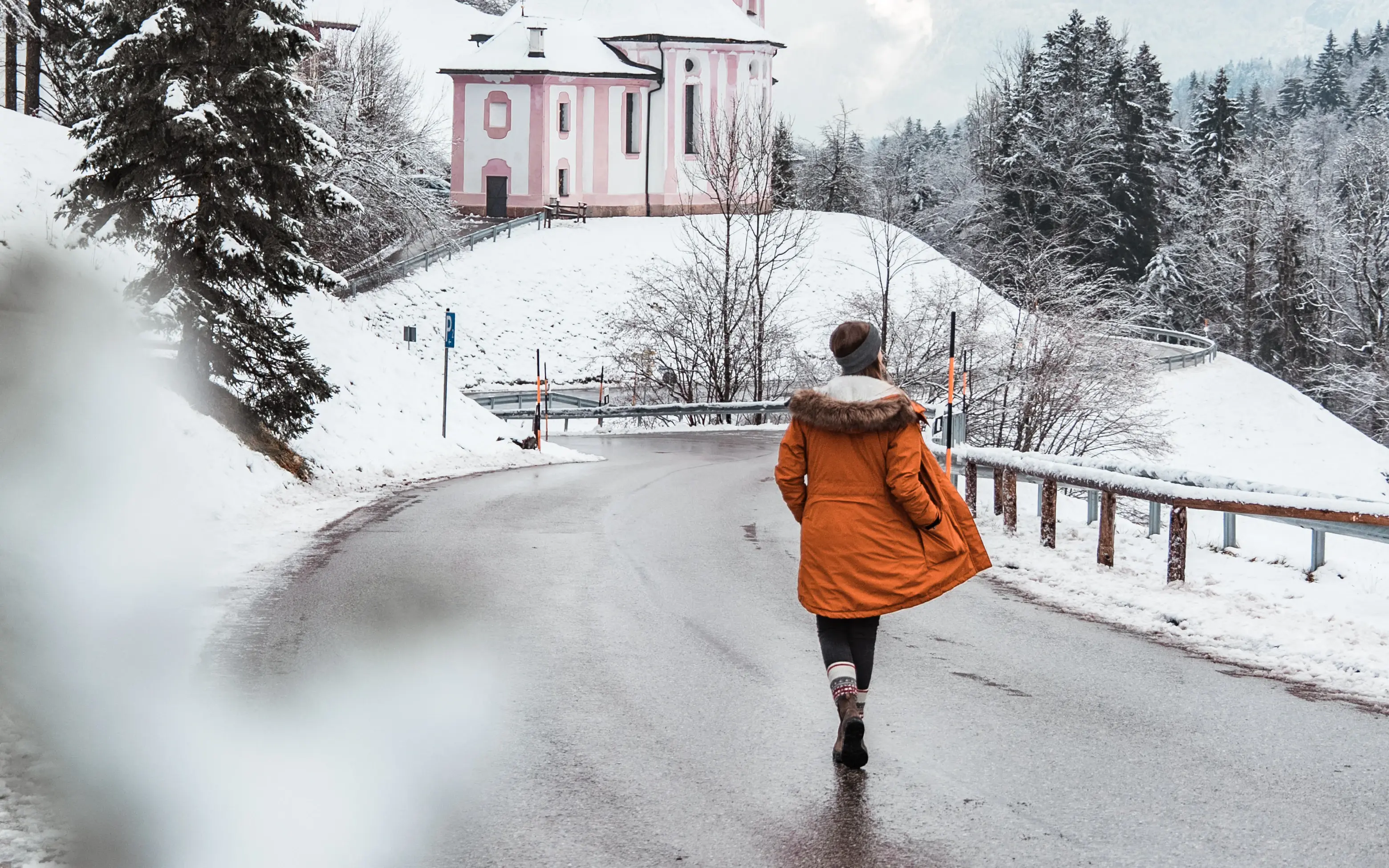 Eine Frau in orangefarbener Winterjacke spaziert eine Straße durch eine schneebedeckte Umgebung entlang.