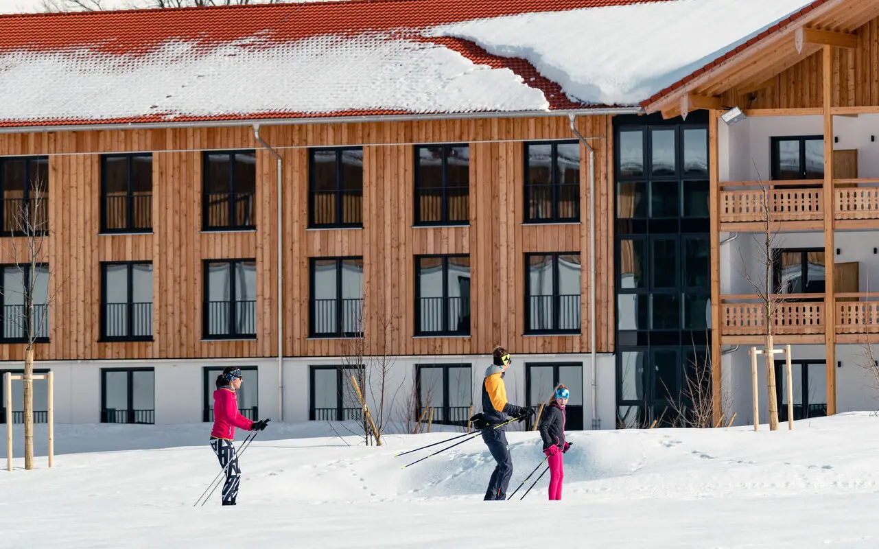 Drei Personen auf Skiern fahren vor einem hölzernen Gebäude durch den Schnee.