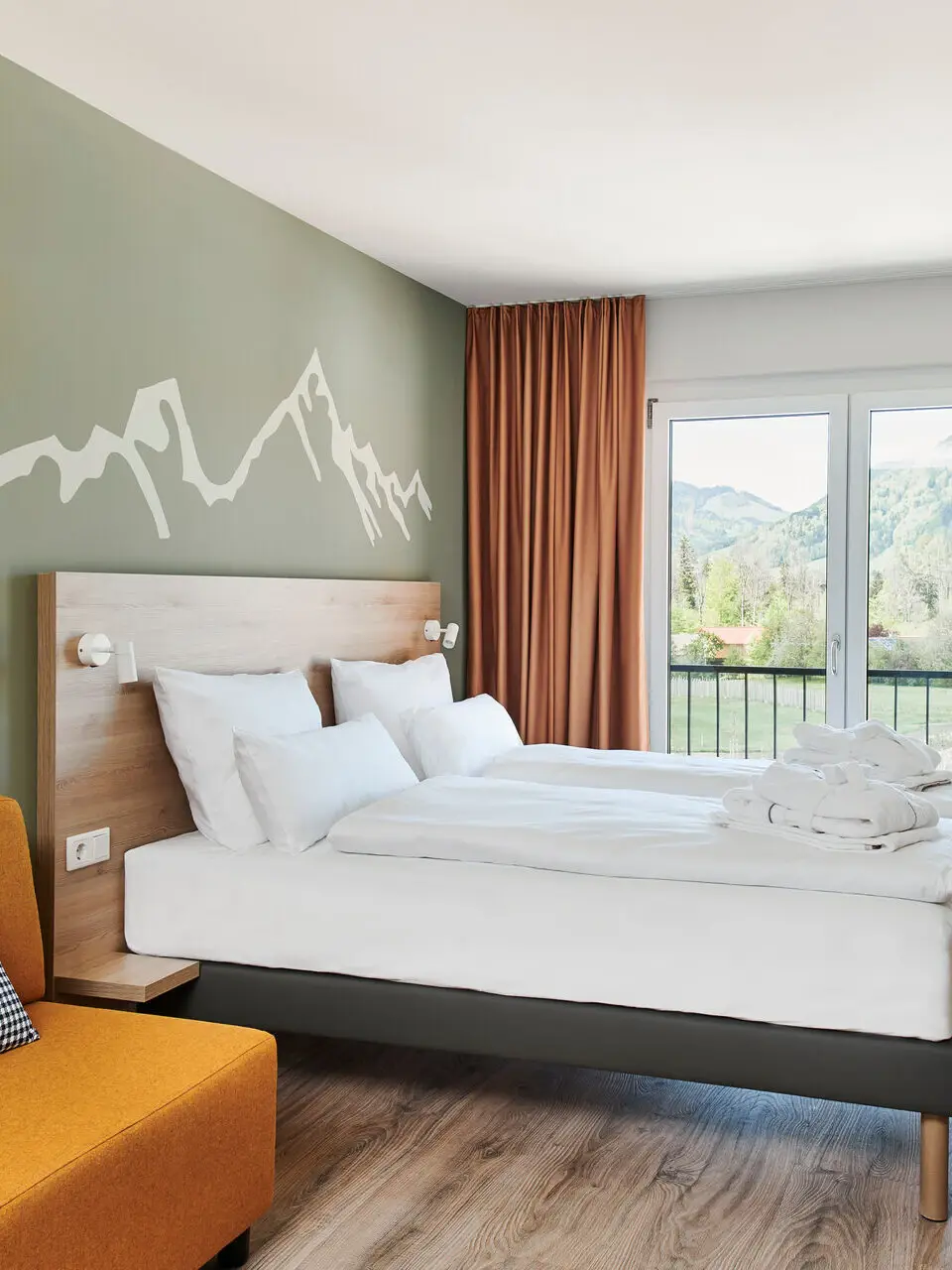 Ein helles Hotelzimmer mit Blick auf Berge und Wälder. Vor einer hellgrünen Wand mit aufgezeichneter Bergkette steht ein Bett mit hölzernem Kopfteil.