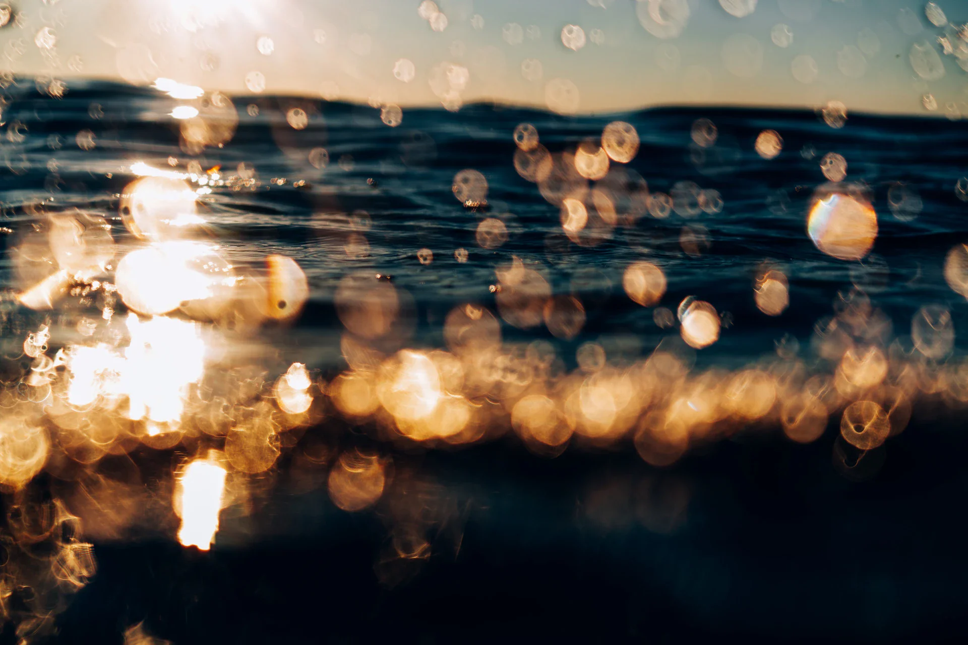 Eine unscharfe Aufnahme von funkelnden Sonnenstrahlen, die sich auf den Wellen des Meeres brechen, und goldenes Licht, das eine warme Atmosphäre schafft.