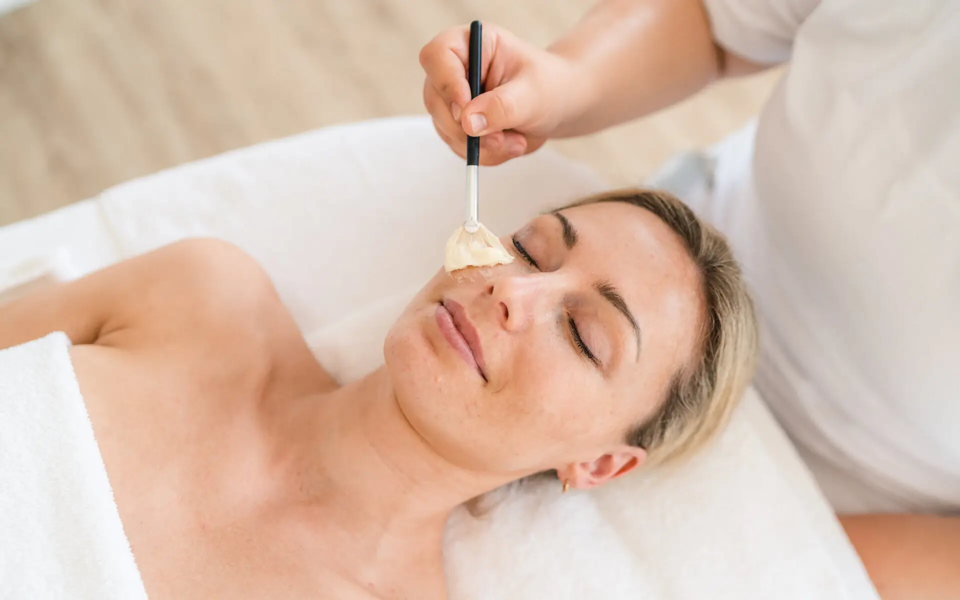 Kosmetik im NIVEA Haus Eine Frau liegt auf dem Rücken und erhält eine Gesichtsanwendung mit einem Kosmetikpinsel. Der Kopf der Frau liegt entspannt ab, das Gesicht ist zur Decke gerichtet. Die Augen der Frau sind geschlossen, sie lächelt leicht und ihre Gesichtszüge wirken entspannt.