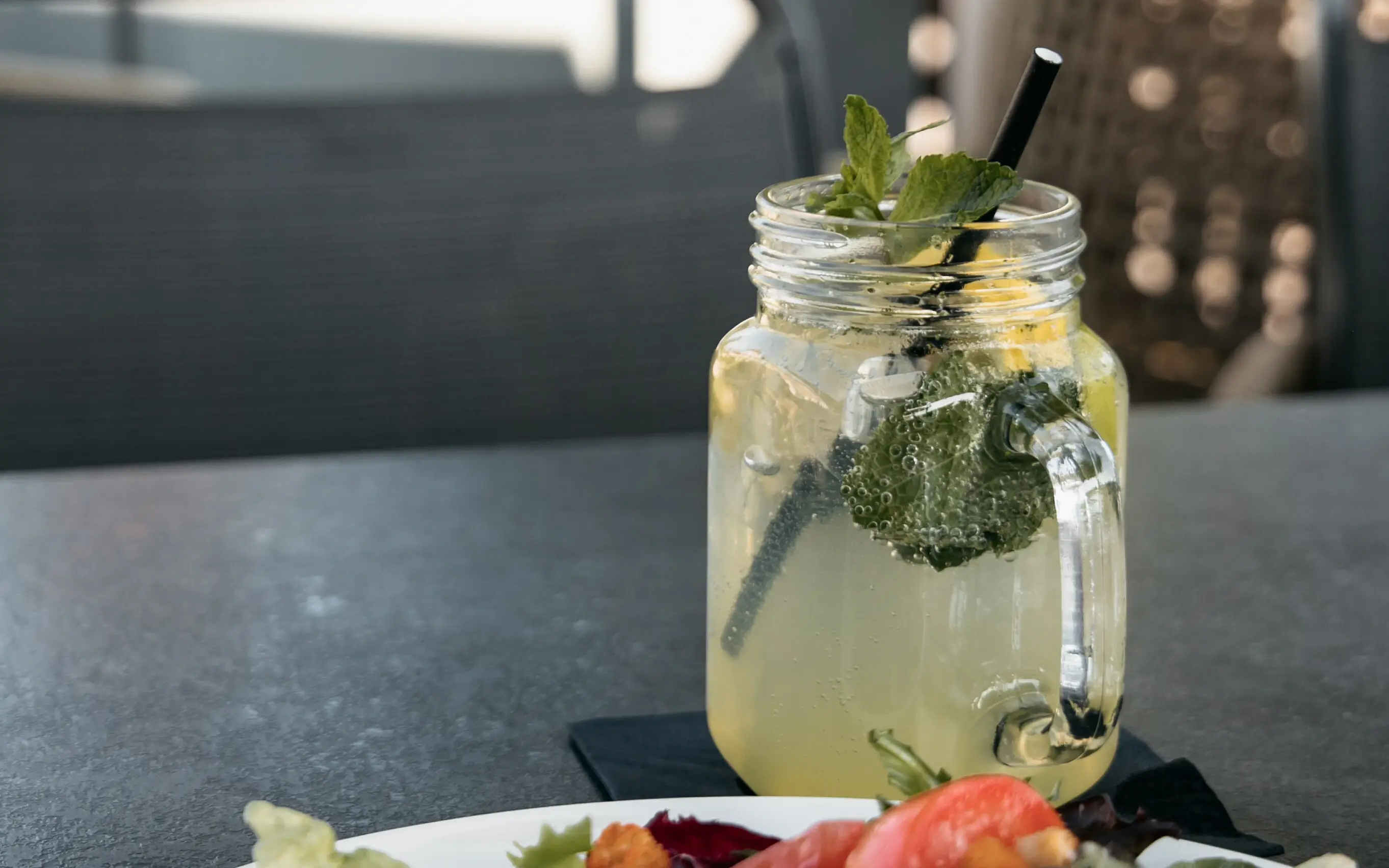 Limonade Auf einem dunklen Tisch steht ein Glas Limonade mit Zitronenscheiben, Minzblättern und einem Strohhalm neben einem zum Teil sichtbaren Salatteller.