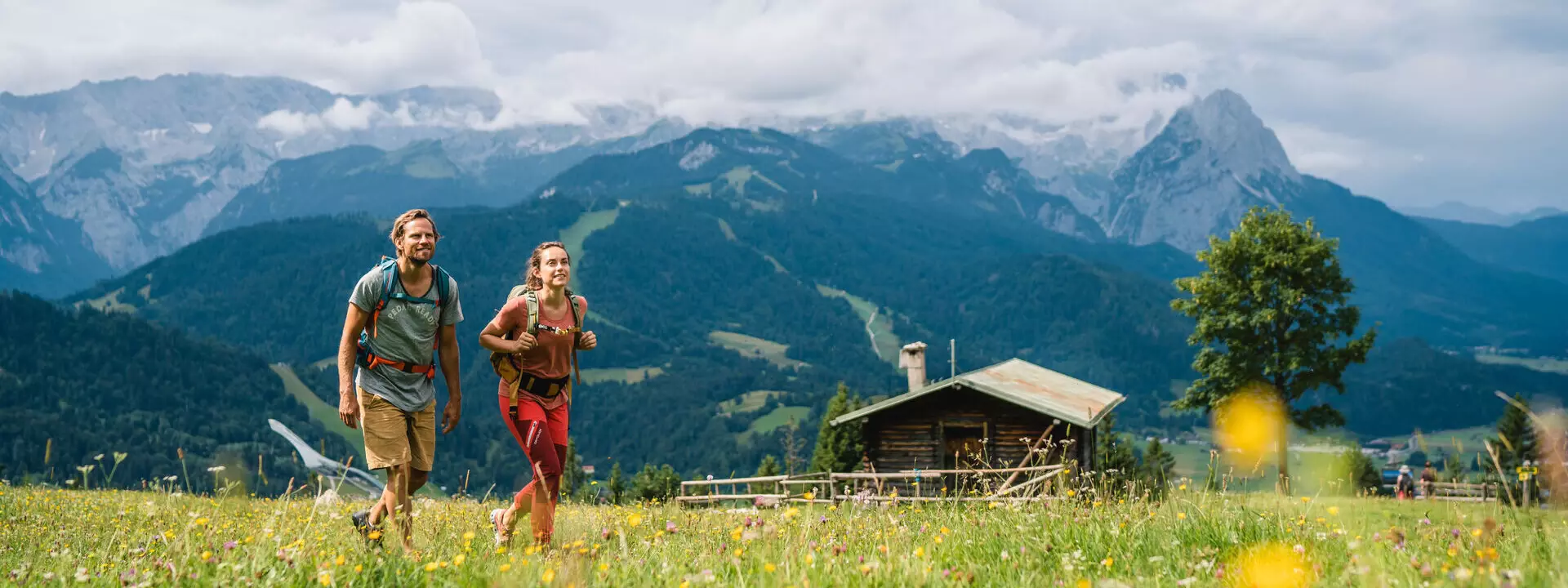 Wandern Garmisch-Partenkirchen Ein Mann und eine Frau wandern durch ein Feld mit Bergen im Hintergrund.